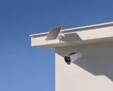 En eufycam 2 pro test visar en vit övervakningskamera med solpanel monterad på en vit vägg under en blå himmel.