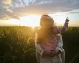 Bambina aggrappata alla schiena di un adulto, con il sole al tramonto che illumina un campo. Un'immagine di puro attaccamento affettivo.