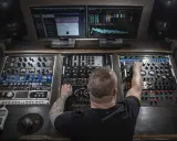 Un homme aux bras tatoués ajuste les réglages d'une console de mixage audio, entouré d'écrans affichant des formes d'ondes et des analyseurs.