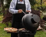 Un homme en tablier noir prépare une pizza et des steaks sur un kamado, un barbecue céramique polyvalent, dans un jardin verdoyant.