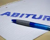 Blauer Stift liegt auf kariertem Papier, auf dem das Wort "Abitur" geschrieben ist. Der **Abitur**-Abschluss rückt näher.