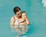 Mama trzyma roczne dziecko na basenie. Maluch bawi się pomarańczową piłeczką.