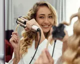 Uśmiechnięta blondynka w szlafroku tworzy piękne fale na włosach za pomocą karbownicy.