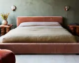 Cama king size tapizada en terciopelo rosa, con mesitas de noche de madera y lámparas de pared.