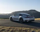 Silberner Porsche 911 Turbo S, ein Sondermodell 2023, parkt auf einer Bergstraße mit sanften Hügeln im Hintergrund.