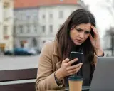 Zmartwiona kobieta czyta na telefonie coś, co wygląda na zniesławienie. Trzyma się za głowę, siedząc przy stoliku z laptopem i kawą.