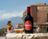 Bottiglia e bicchiere di Amara, un amaro che celebra come l'arancio amaro trama i sapori siciliani. Sfondo sfocato di un borgo.