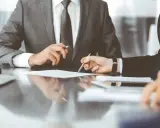 Dwóch mężczyzn w garniturach przy biurku omawia dokumenty. Jeden z nich wskazuje piórem na papier, wyjaśniając, kto to syndyk.