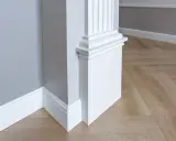 Biały pilaster z żłobieniami i elegancką bazą, idealny do wnętrzarskiego bloga. Drewniana podłoga w jodełkę i szara ściana tworzą stylowe tło.