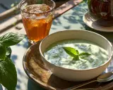 Deliciosa crema de pepino servida en un cuenco, acompañada de un vaso de té helado y hojas verdes frescas.