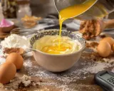Préparation d'une pâte à bombe : du beurre fondu est versé dans un bol de crème. Farine, cacao, œufs et thermomètre à proximité.