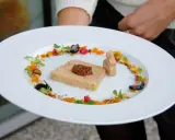 Une présentation raffinée de foie gras en entrée, ornée de fruits secs, de baies et de pousses vertes sur une assiette blanche texturée.