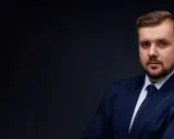 Mężczyzna w granatowym garniturze i krawacie, z zarostem, na ciemnym tle.