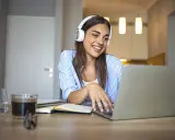 Uśmiechnięta studentka w słuchawkach pracuje przy laptopie. Czy muzyka pomaga w nauce? Ona chyba tak, bo wygląda na zrelaksowaną i skupioną.