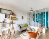 Idée aménagement studio 25m2 : un espace salon cosy avec canapé, table haute et étagères murales, séparé d'une chambre par une bibliothèque ajourée.