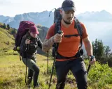 Dos excursionistas con mochilas y bastones caminan por una ladera montañosa.