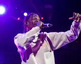 Travis Scott na scenie, w białej bluzie, z mikrofonem w dłoni. Jego energia inspiruje wielu **współautorów piosenek**.