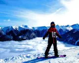 Snowboardzista w kurtce hardshell stoi na szczycie góry, nad morzem chmur. Góry w tle.