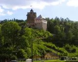 Zamek w Czchowie góruje nad zielonym wzgórzem, otoczony drzewami. Na szczycie wieży powiewa flaga.