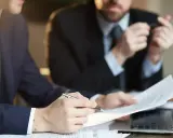 Dwóch mężczyzn w garniturach analizuje dokumenty. Jeden z nich wskazuje piórem na umowę, która może być naganą dla pracownika.