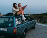 Dwie dziewczyny siedzą na dachu niebieskiego kombi, podziwiając widok na port. To idealny moment na podróż samochodem po Holandii.