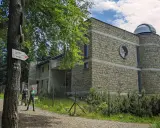 Kamienny budynek obserwatorium na szczycie Lubomir. Dwoje turystów podziwia widok, a obok drzewa i szlak PTTK.