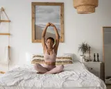 Frau praktiziert Yoga im Bett, mit geschlossenen Augen und erhobenen Armen. Ein Bild von einem Strand hängt an der Wand.