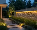 Allée de dalles éclairée par des spots au sol, bordée d'herbes hautes et d'un mur de pierre illuminé par une lumière 4000k couleur.
