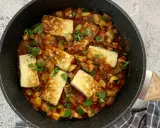 Smażony ser halloumi z warzywami w sosie pomidorowym, posypany natką pietruszki, w czarnej patelni.