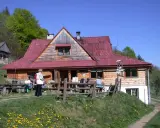 Schronisko pod Bereśnikiem, drewniany budynek z czerwoną dachem, otoczony zielenią. Kilka osób odpoczywa na ławkach przed schroniskiem.