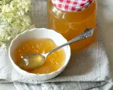 Ein Löffel voll goldgelbem Holunder Apfel Gelee in einer Schale, daneben ein Glas mit dem Gelee und Holunderblüten.