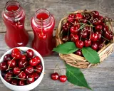 Dwa szklane dzbanki z rubinowym sokiem z czereśni, obok koszyk pełen świeżych owoców i biała miseczka z czereśniami.