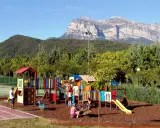 Parque infantil colorido en un camping de Huesca, con niños jugando y montañas imponentes al fondo.