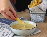 Une frite trempée dans une sauce moutarde miel onctueuse. Des frites dorées dans un panier en métal à côté.