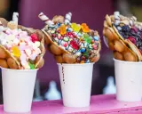 Trois bubble waffles colorés, garnis de bonbons, guimauves et fruits, présentés dans des gobelets blancs.