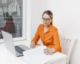Kobieta w okularach i pomarańczowej bluzce analizuje dokumenty przy laptopie. Dowiaduje się, jak zostać notariuszem.