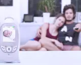 Ein Babyphone mit Bildschirm zeigt ein Baby. Im Hintergrund entspannen sich Eltern auf dem Sofa.