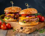 Dwa soczyste burgery z serem, warzywami i piklami na drewnianej desce, obok pomidorków koktajlowych.