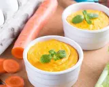 Dwa białe naczynia z pomarańczowym puree z marchewki, udekorowane miętą. Obok marchewka, jajka i seler.