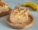 Pyszny chlebek bananowy fit z płatkami owsianymi na wierzchu, podany na drewnianym talerzyku. Obok leży dojrzały banan.