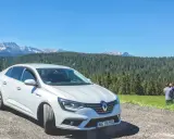 Biały Renault Megane Sedan zaparkowany na szutrowej drodze z widokiem na góry i las. Na trawie stoi mężczyzna z dzieckiem.