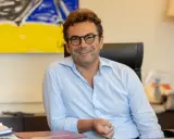 A man with glasses and a light blue shirt smiles while sitting at a desk with abstract art in the background.