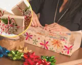 Osoba pakuje prezent w kolorowy papier. W tle widać kilka prezentów w brązowym papierze.