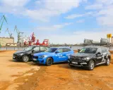 Trzy SUV-y BMW stoją na placu budowy, w tle portowe dźwigi i budynki.