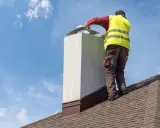 Pracownik na dachu montuje przedłużenie komina. Niebo błękitne z chmurkami.