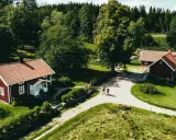 Två röda hus med tegeltak vid Rya Åsar, Borås. En grusväg leder förbi husen genom ett grönskande landskap med skog och ängar.