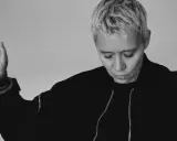 Jennifer Cardini, con i suoi capelli biondi corti e un'espressione pensierosa, alza un braccio con un gesto delicato. Indossa una giacca scura con zip.