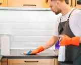 Hombre con guantes naranjas desinfecta la cocina, rociando la encimera con un spray limpiador.