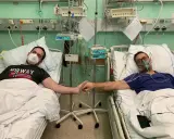 Dwóch mężczyzn w szpitalnych łóżkach, po operacji guza nerki, trzyma się za ręce. Jeden ma maskę tlenową, drugi zakrywa usta.