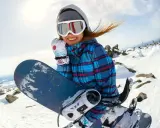 Uśmiechnięta snowboardzistka w goglach i czapce trzyma deskę snowboardową na tle zaśnieżonych gór.